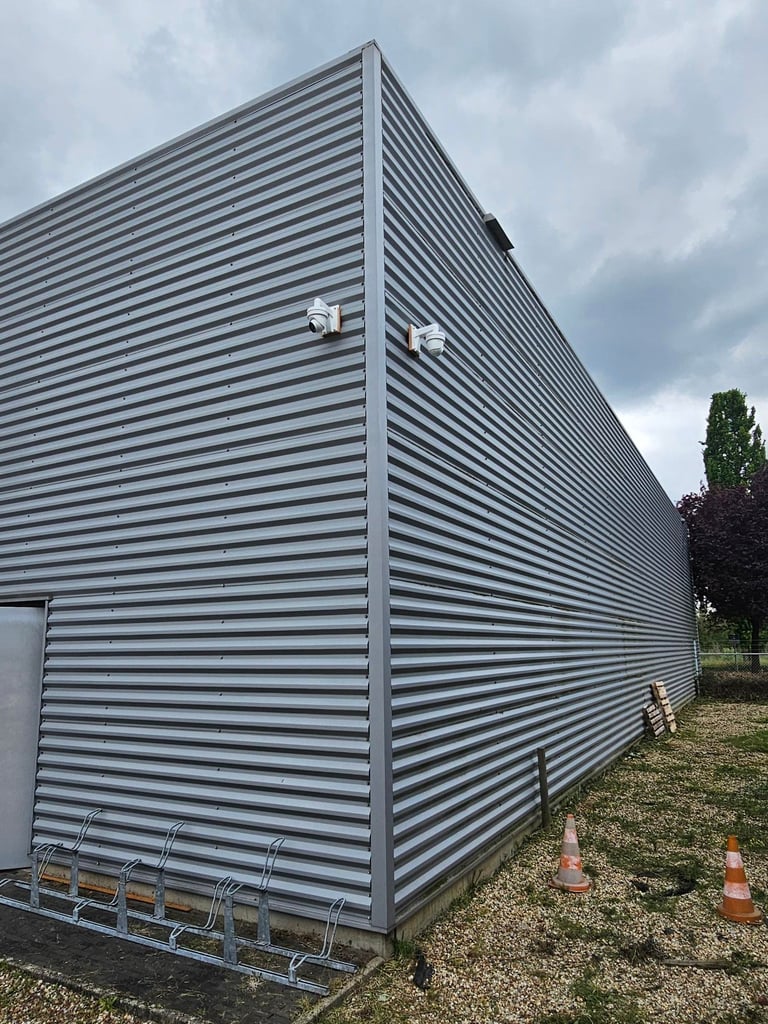 Modern gray corrugated metal building exterior with horizontal ridged panels, security lights, and traffic cones on ground